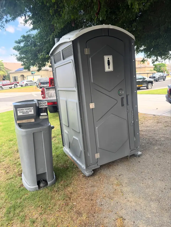 Gray standard porta potty with a two-sink handwash station on a lawn